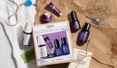 Flat lay of Kiehl's skincare products displayed on crumpled brown paper. The arrangement includes various bottles and jars, featuring a prominent box labeled "Radiant Skin Recovery," showcasing the products inside.