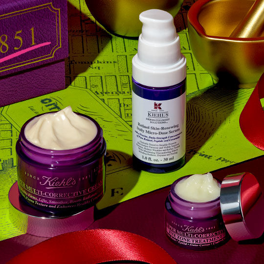 Tre Kiehl's Super Multi-Corrective produkter – en kremkrukke, en øyekrem og en serumflaske – er arrangert på et rødt bånd, ved siden av en lilla eske og gullfargede festdekorasjoner, mot en limegrønn bakgrunn med et kartmønster.