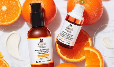 Two Kiehl's skincare products are displayed on a surface with halved oranges and cream swirls. The products include a Powerful-Strength Line-Reducing Concentrate and an Eye Serum, both prominently labeled.