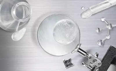 A magnifying glass focuses on a droplet of water on a metallic surface, surrounded by a glass vial, a test tube, and molecular models.