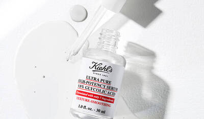 A clear bottle of Kiehl's Ultra Pure High-Potency Serum with 38% Glycolic Acid, partially spilled, on a light background. The bottle is labeled with product information, emphasizing texture-smoothing benefits.