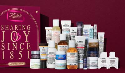 Kiehl's Adventskalender gaveeske i dyp rød med gulltekst, vist ved siden av 24 mini hudpleieprodukter inkludert Ultra Facial Cream, Calendula Toner, Retinol Serum og Lip Balm #1. En rosa spraymalt grafikk på siden fremhever verdien av settet.