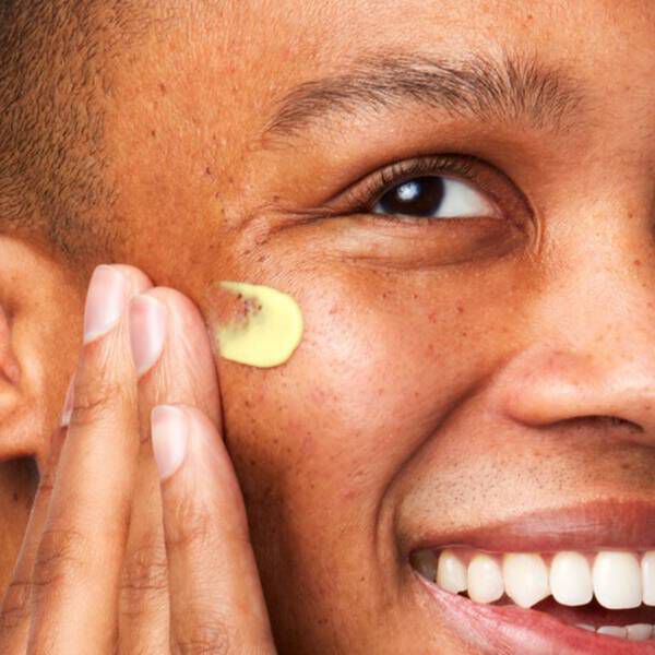 A person smiling while applying a small amount of skincare product to their cheek with one hand.