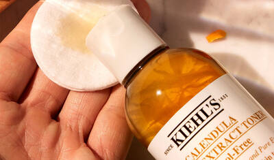 A hand holding a cotton pad with a bottle of Kiehl's Calendula Extract Toner in the background. The toner is a golden yellow and labeled with the brand name.