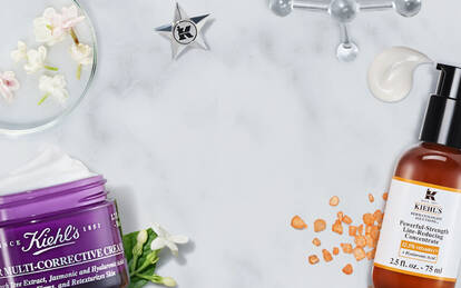 Produkter fra Kiehl's plassert på en marmoroverflate, inkludert en serumflaske, en fuktighetskrem i en lilla krukke, og en liten flaske med behandling. Blomster og andre detaljer er også synlige.