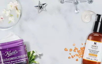 Produkter fra Kiehl's plassert på en marmoroverflate, inkludert en serumflaske, en fuktighetskrem i en lilla krukke, og en liten flaske med behandling. Blomster og andre detaljer er også synlige.