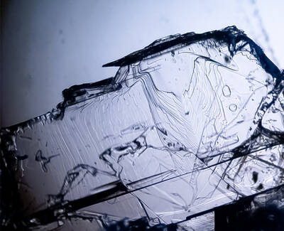 Close-up image of a translucent crystal fragment with intricate surface textures and patterns, exhibiting light reflections and refractions against a soft background.