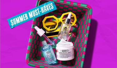 Et lilla banner med en grønn og rosa flettet kurv fylt med Kiehl's hudpleieprodukter og gule solbriller, med blå teip der det står 'SUMMER MUST-HAVES'.