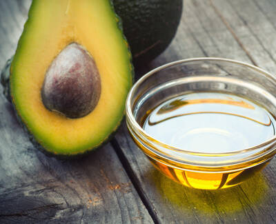 A halved avocado with a seed next to a small glass bowl of golden oil, placed on a rustic wooden surface.