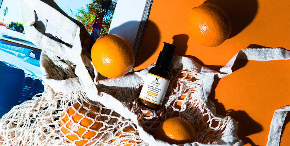 A flat lay featuring a bottle of skincare serum nestled in a cream-colored net bag, surrounded by bright oranges on an orange background, with a magazine featuring a car in the corner.
