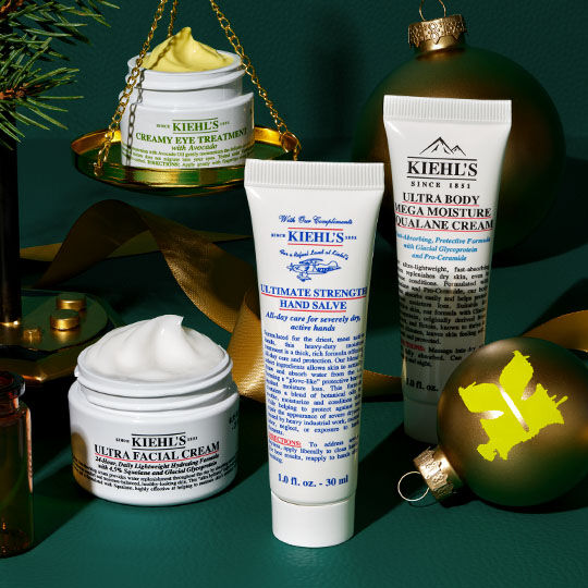 Et festlig arrangement av Kiehl's produkter, inkludert Creamy Eye Treatment på gylne skjell, Ultra Facial Cream, Ultimate Strength Hand Salve og Ultra Body Mega Moisture Squalane Cream, omkranset av grønne ornamenter, grantraer og gullbånd på en mørkegrønn bakgrunn.