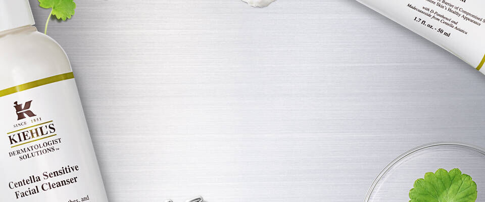 Bilde av Kiehl's produkter, inkludert en ansiktsrens og krem, plassert på en metalloverflate med urter og laboratorieutstyr i bakgrunnen.