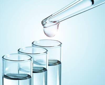 A dropper is dispensing a droplet of liquid into one of three test tubes filled with clear liquid.