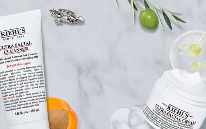 Bildet viser Kiehl's hudpleieprodukter, inkludert rensegel, toner og fuktighetskrem, plassert på en marmoroverflate sammen med noen naturlige elementer som oliven og en mandarin.