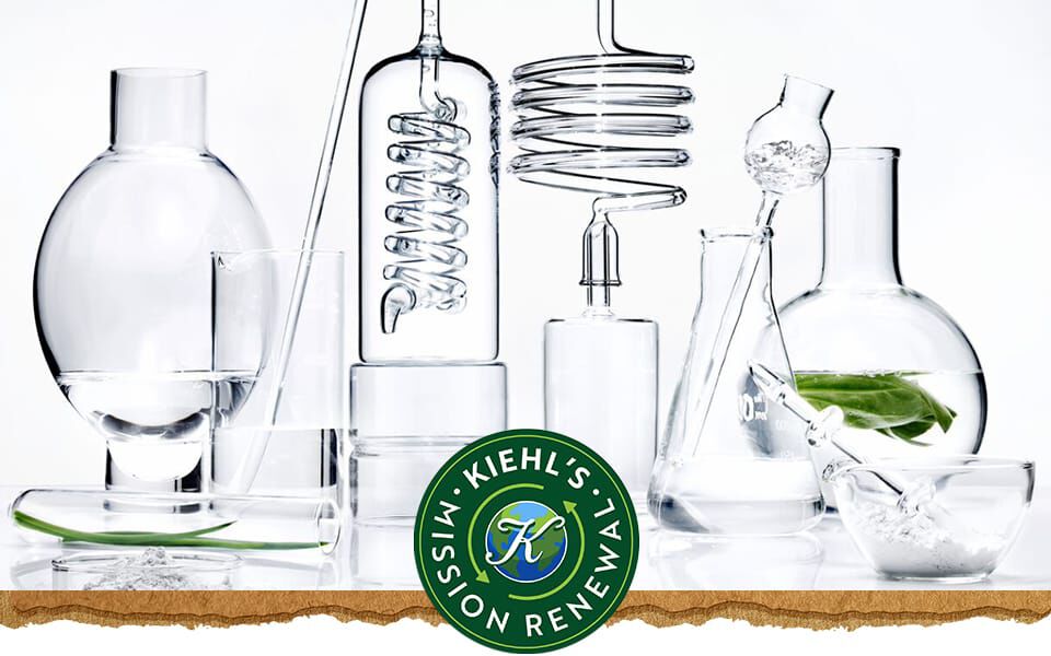 En samling laboratoriekar i klart glass fylt med vann og et grønt blad, med Kiehl's logo til høyre.