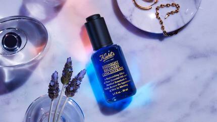 A blue bottle of Kiehl's Midnight Recovery Concentrate displayed on a marble surface, surrounded by lavender flowers and gold jewelry, with soft lighting creating a calming atmosphere.