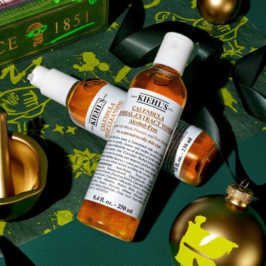 Kiehl’s Calendula Herbal-Extract Toner og serum arrangert med gyldne juleornamenter og grønt bånd på en festlig mørkegrønn bakgrunn.