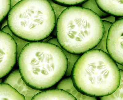 A close-up view of sliced cucumbers, showcasing their vibrant green skin and light-colored flesh with visible seeds. The slices are arranged closely together.