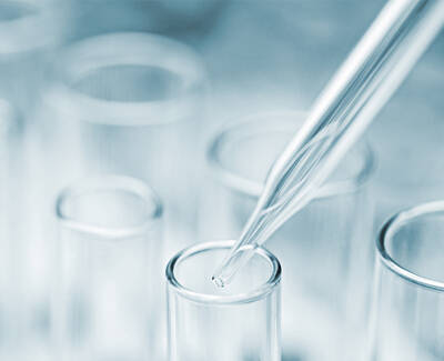 A close-up of a dropper poised over a clear test tube, with a blurred background of additional test tubes. The image conveys a laboratory setting focused on precise measurements.