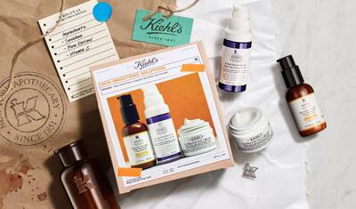 A flat lay of Kiehl's skincare products on a tan surface, including a cardboard box featuring "Skin Smoothing Solutions" and various bottles of serums and creams. There are also tags and a blue note in the background.