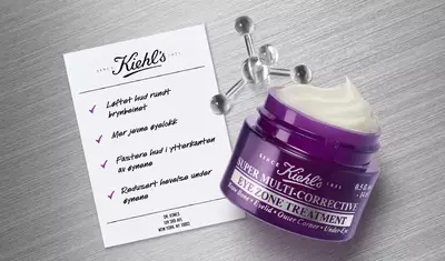 A jar of Kiehl's Super Multi-Corrective Eye Zone Treatment beside a printed list of benefits, highlighting improvements in brow area lift, eyelid smoothing, outer corner firmness, and under-eye de-puffing percentages.