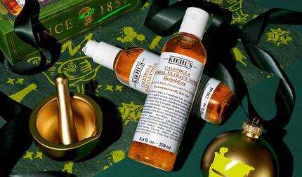 På en festlig, mørkegrønn bakgrunn med gullfargede aksenter ser du to flasker: "Kiehl's Calendula Deep Cleansing Foaming Face Wash" og "Kiehl's Calendula Herbal-Extract Toner", begge med gjennomsiktig emballasje og ravfarget vaeske. Scenen inkluderer en morter og støter i gull, et gullornament med en grønn "K"-logo, svart bånd, og bakgrunnen er dekorert med gullillustrasjoner.