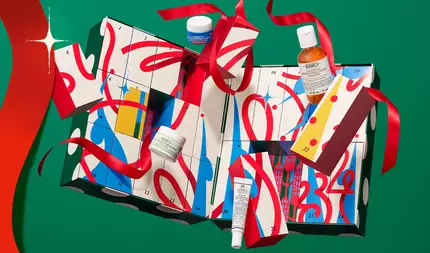 Kiehls jule-adventskalender med nummererte gaveesker og hudpleieprodukter, pakket inn i rødt bånd på en festlig grønn bakgrunn.