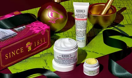 En festlig grafikk som viser en hvit tube med "KIEHL'S ULTRA FACIAL BARRIER-HYDRATING CLEANSER", en hvit krukke med "KIEHL'S ULTRA FACIAL CREAM" med synlig hvit krem, og en liten hvit krukke med "KIEHL'S CREAMY EYE TREATMENT WITH AVOCADO" med synlig blekgul krem, alt arrangert på en mørkerød og grønn bakgrunn med kartmønster. En morter og mortel i gull er synlig øverst til høyre. Øverst til venstre ses en julekule i gull med rosa fuglemønster og en rød gaveeske med "SINCE 1851" og hodeskaldemønster. Rundt produktene er det drapert et sort bånd.